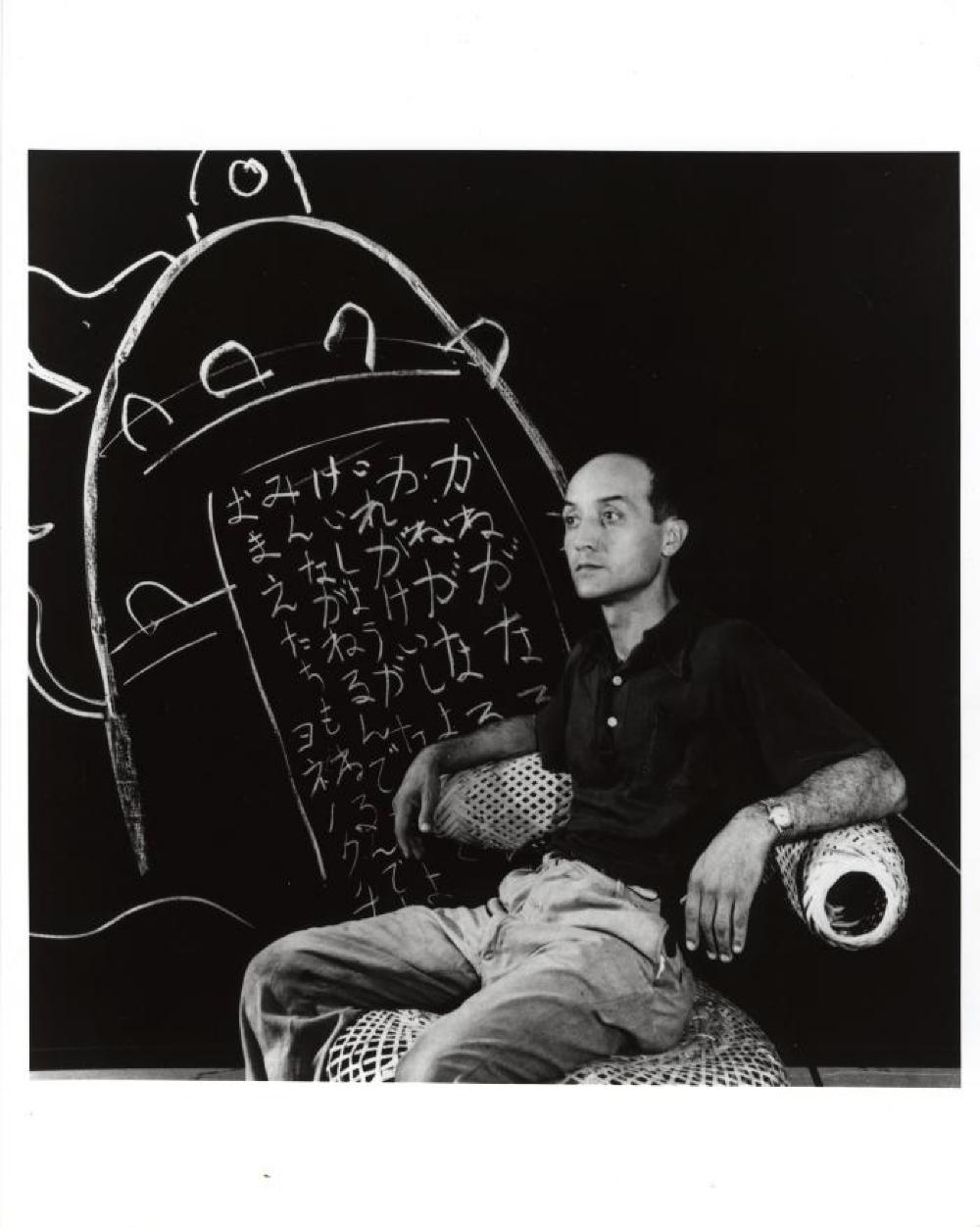 Isamu Noguchi seated in Basket Chair, with writing of his father's poem at the Mitsukoshi Dept Store exhibition (Nihombashi Head Store), Tokyo, Aug. 1950.